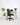 A black ergonomic office chair with a high back and five-star wheeled base, positioned in front of a light wood desk with a laptop and flowers, set against a white wall and floor with potted plants on the left.